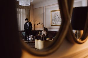 man in black suit standing and a woman sitting on sofa